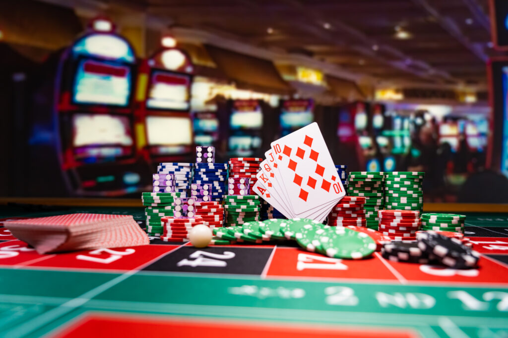 A photo of casino chips, dice and playing cards on top of a roulette table with slot machines in the background.