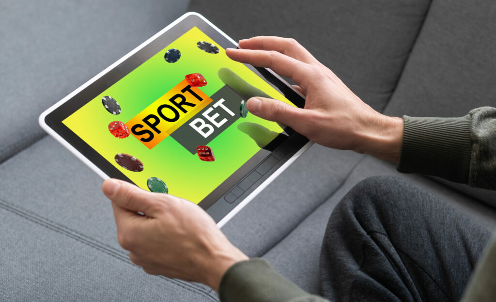 Hands holding a tablet with a screen showing a yellow and green background with "SPORT BET" in the centre. Around this are poker chips and dice.
