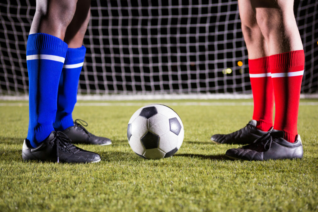 Male players standing by soccer ball against goal post on playing field at night.