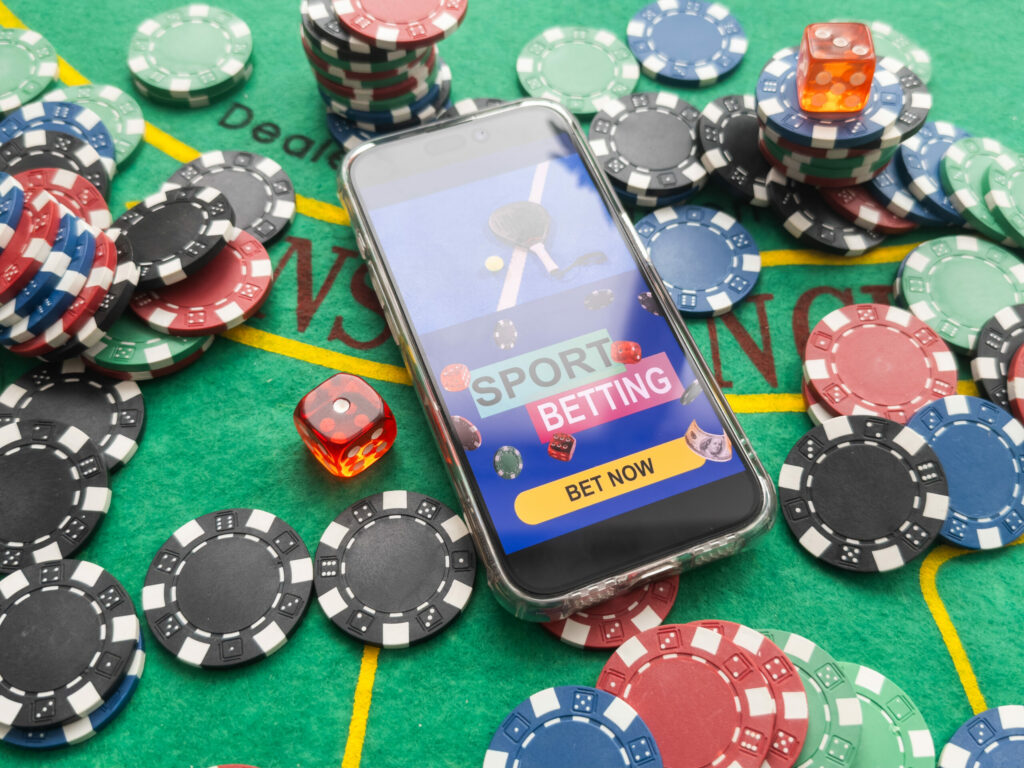 A smartphone on a blackjack table with "SPORT BETTING" on the screen, surrounded by chips, playing cards and dice.