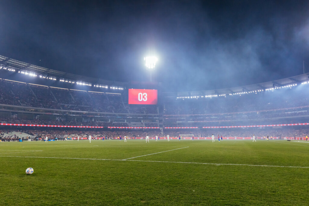 Football match at night in a filled stadium. There is football in the foreground with players in the background, and the full stands of football fans behind them.