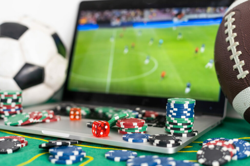 Casino chips and red dice on and beside a laptop on a green table. The screen is showing a football match. To the left of the laptop is a football, and to the right of the laptop is an American football.