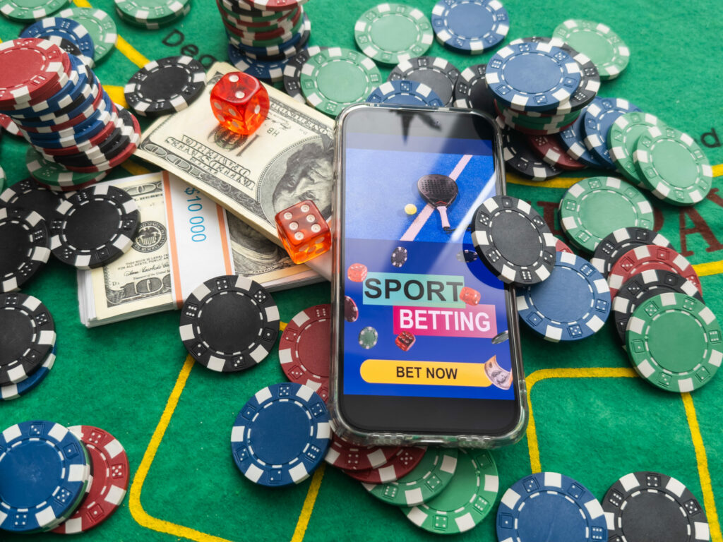 Smartphone on a green table surrounded by casino chips, dice and American dollars. On the smartphone's screen are the words "SPORT BETTING" and "BET NOW" on a blue background with a tennis racket and tennis ball.