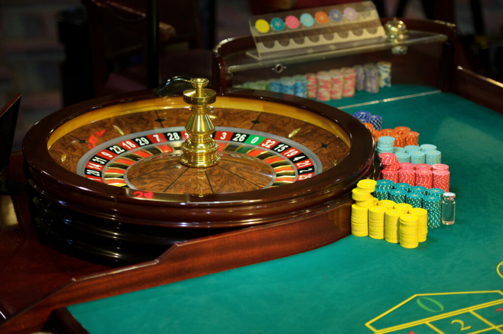 Roulette wheel on a green casino table with organised piles of yellow, green, red, teal/cyan, orange and dark blue chips beside it.