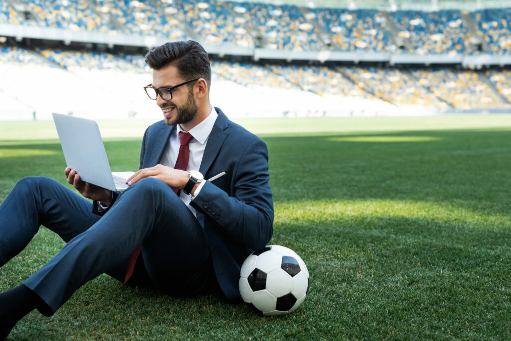 Smiling young businessman in suit with laptop and soccer ball sitting on football pitch at stadium.