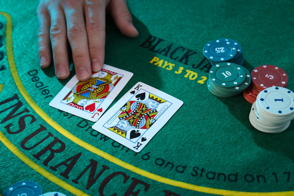 A man, out of frame, placing a Jack of Hearts beside a King of Spades on a green blackjack table. There are also casino chips on the table.