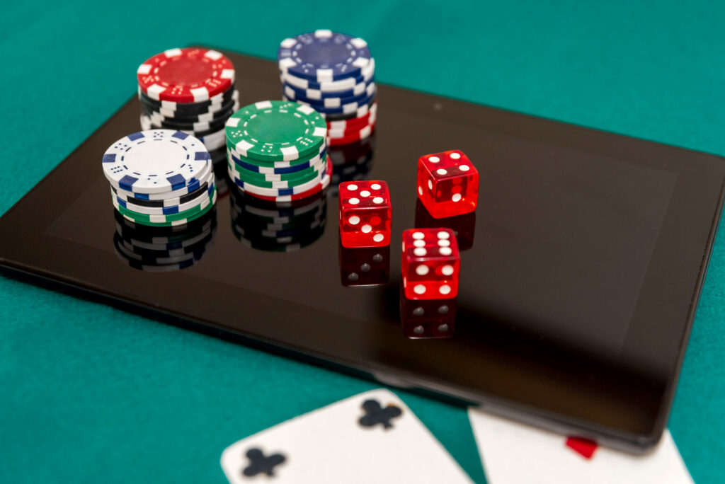 Close up of poker chips and red dice on digital tablet with playing cards on green felt.