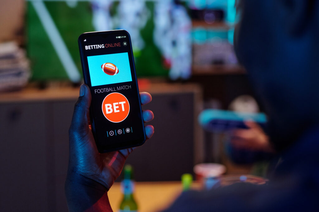 Man holding smartphone displaying online sports betting app with football match bet option visible on screen, sitting indoors with blurred background.