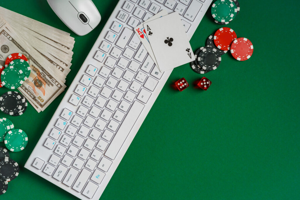 A white keyboard and mouse on a green felt casino table. On the keyboard are two playing cards - the King of Hearts and Ace of Clubs. Beside the keyboard and mouse are a stack of 50 dollar bills, casino chips and dice.